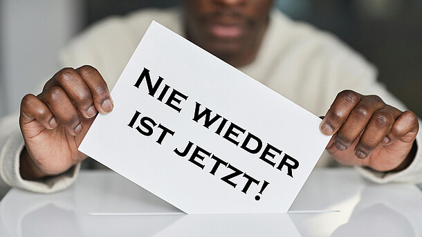 Eine Person wirft einen Zettel mit der Aufschrift "Nie wieder ist jetzt" in eine Wahlurne